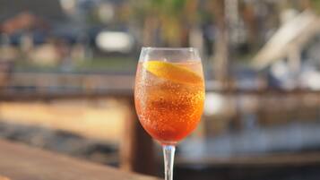 A glass of red beverage with an orange slice on a wooden table with a marina and palm trees in the background.