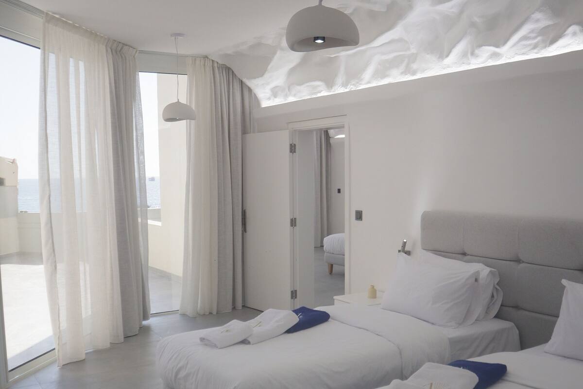 A modern hotel room with two beds, a large window with sheer curtains, and a view of the sea.