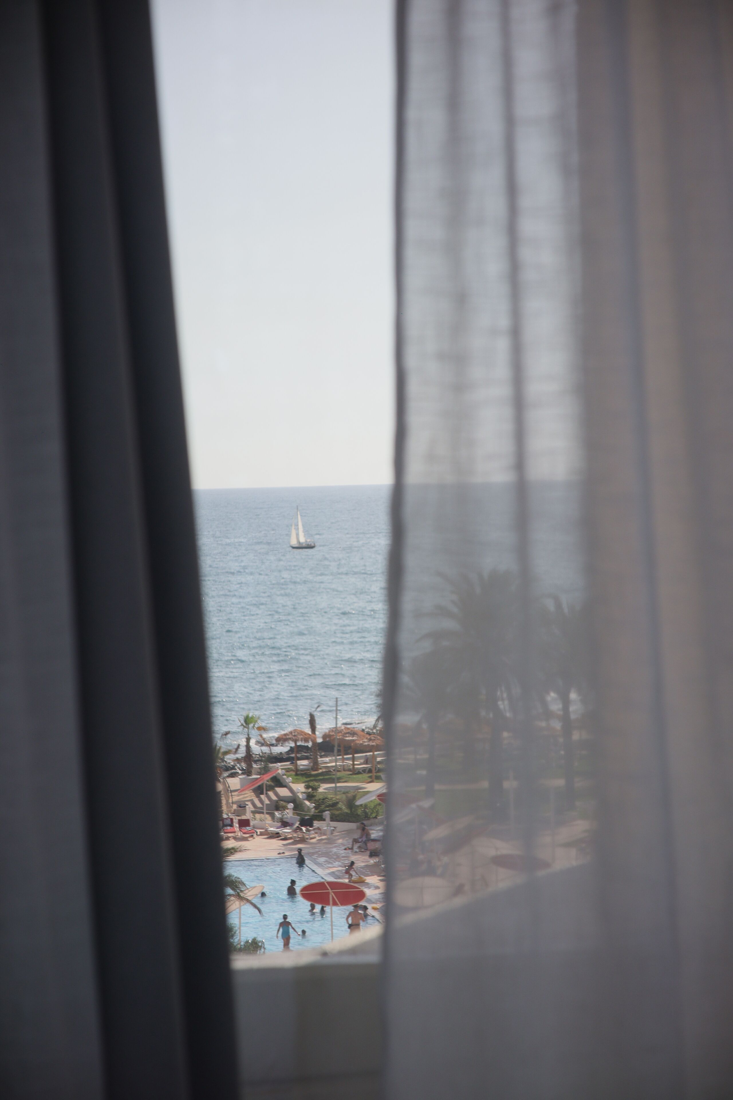 A view through a window with sheer curtains, featuring a beach with a sailboat on the water and a pool area with umbrellas and people.