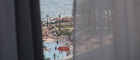 A view through a window with sheer curtains, featuring a beach with a sailboat on the water and a pool area with umbrellas and people.