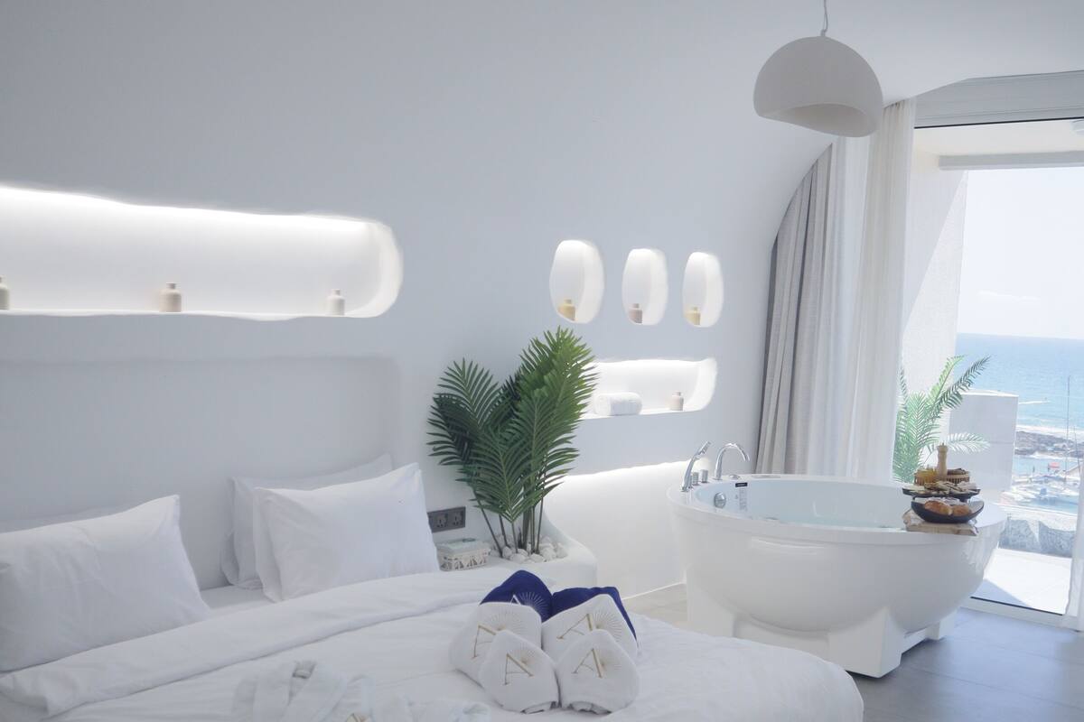 A modern bedroom with a large bed, a bathtub, and a view of the ocean.