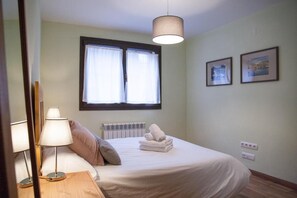 Un dormitorio con cama, lámparas de noche, ventana con cortinas, radiador y dos cuadros en la pared.