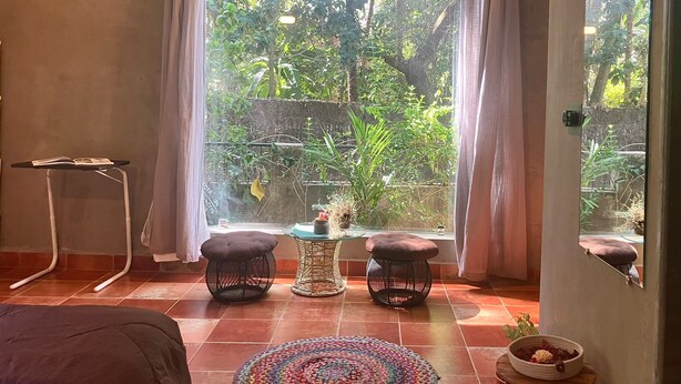 A room with a large window, two black stools, a small table, and a colorful rug. - Casa ruya-Tierra (Benaulim)