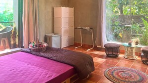 A bedroom with a bed, a small table, a chair, a storage unit, and a view of greenery outside. - Casa ruya-Tierra (Benaulim)