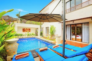 A swimming pool with blue lounge chairs and an umbrella, surrounded by plants and a building with large windows. - Villa Alessandra Seminyak (Bali)