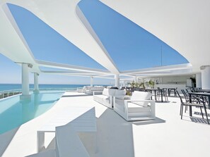 Un área lounge moderna al aire libre con piscina, mobiliario blanco y vista despejada al océano.
