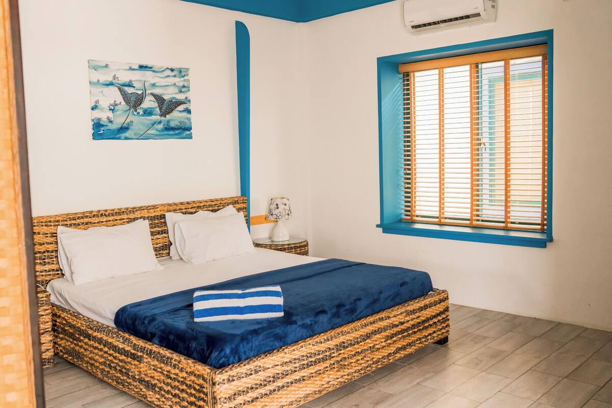 A bedroom with a wicker bed, a window with blinds, a wall-mounted air conditioner, and a painting of birds.