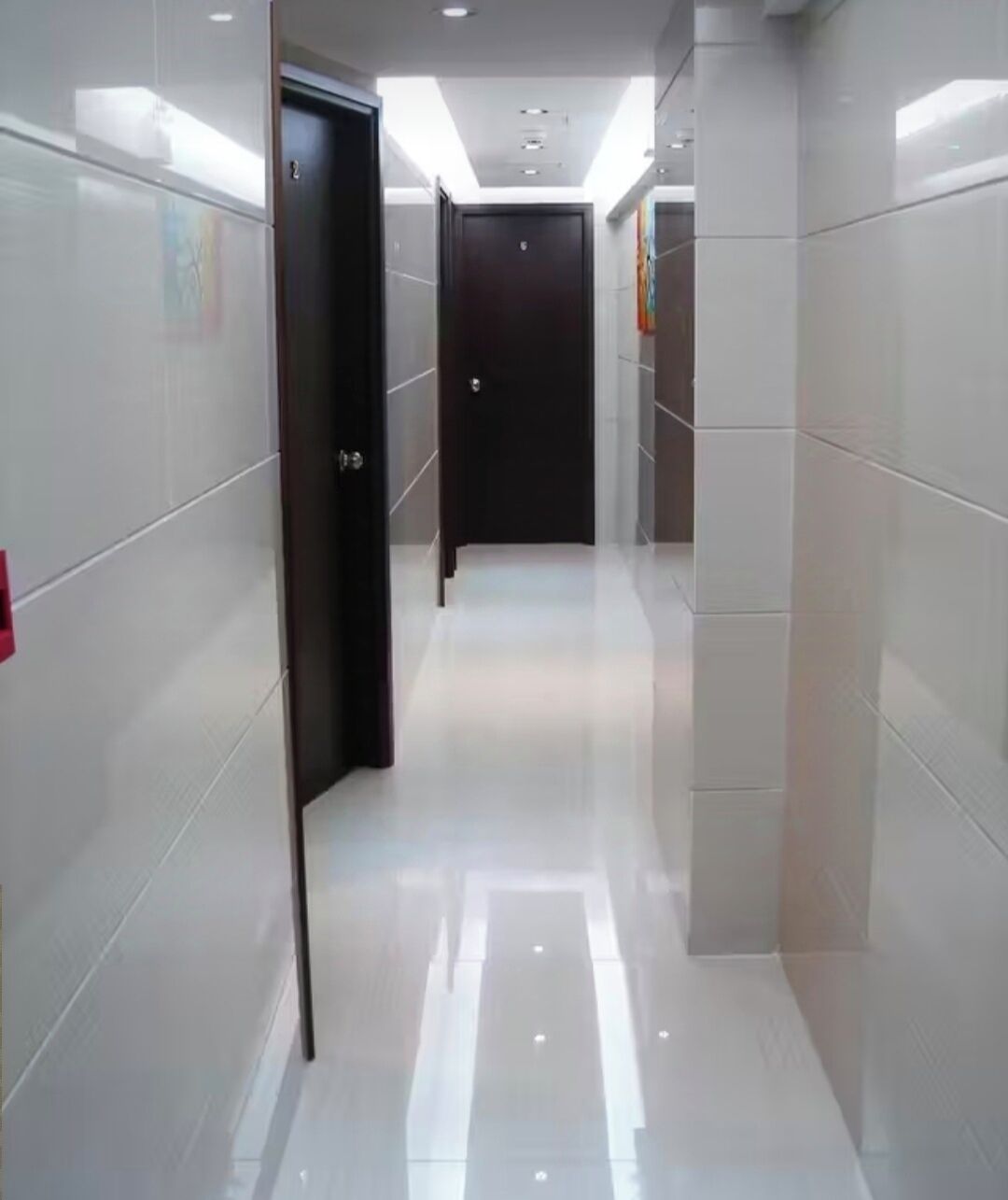 A corridor with white tiled walls and a shiny floor, leading to a dark door at the end.