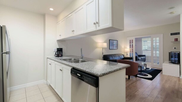 Una cocina moderna con armarios blancos, lavavajillas y cafetera. Hay una zona de comedor con sofá y televisor.