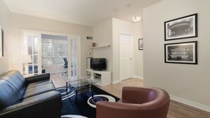 Una sala de estar moderna con un sofá de cuero, una mesa de centro de vidrio, un sillón marrón y un televisor sobre un mueble blanco. Hay cuadros enmarcados en la pared y una puerta que da a otra habitación.