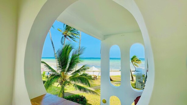 Vista a través de un arco blanco hacia una playa con palmeras y un cielo azul despejado.