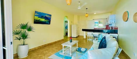 A spacious living area with a sofa, a coffee table, and a potted plant. There is a kitchen area with a refrigerator and cabinets.