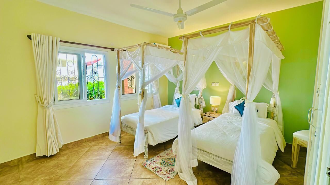 A room with four beds, each with a canopy, a window with curtains, a ceiling fan, and a small table with a lamp.