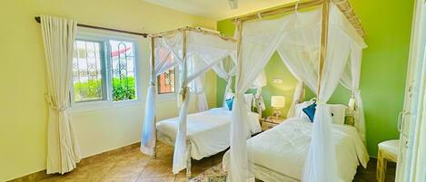 A room with four beds, each with a canopy, a window with curtains, a ceiling fan, and a small table with a lamp.
