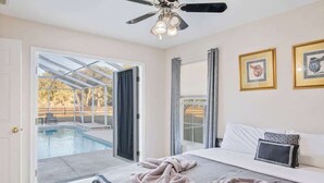 Un dormitorio con cama, ventilador de techo, ventana grande y vista a una piscina al aire libre.