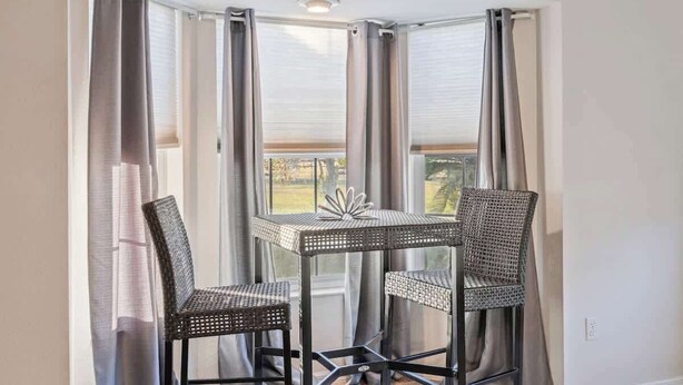Habitación de esquina con una mesa y dos sillas, ventanas amplias con cortinas y vistas al exterior.
