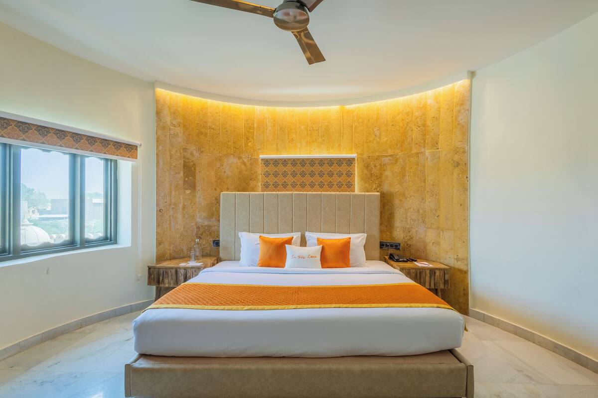A modern hotel room with a large bed, a ceiling fan, a nightstand, and a view of the city through the window.