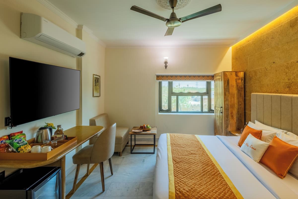 A hotel room with a bed, a desk with a TV, a chair, a small table with a lamp, and a window with a view of the outside.