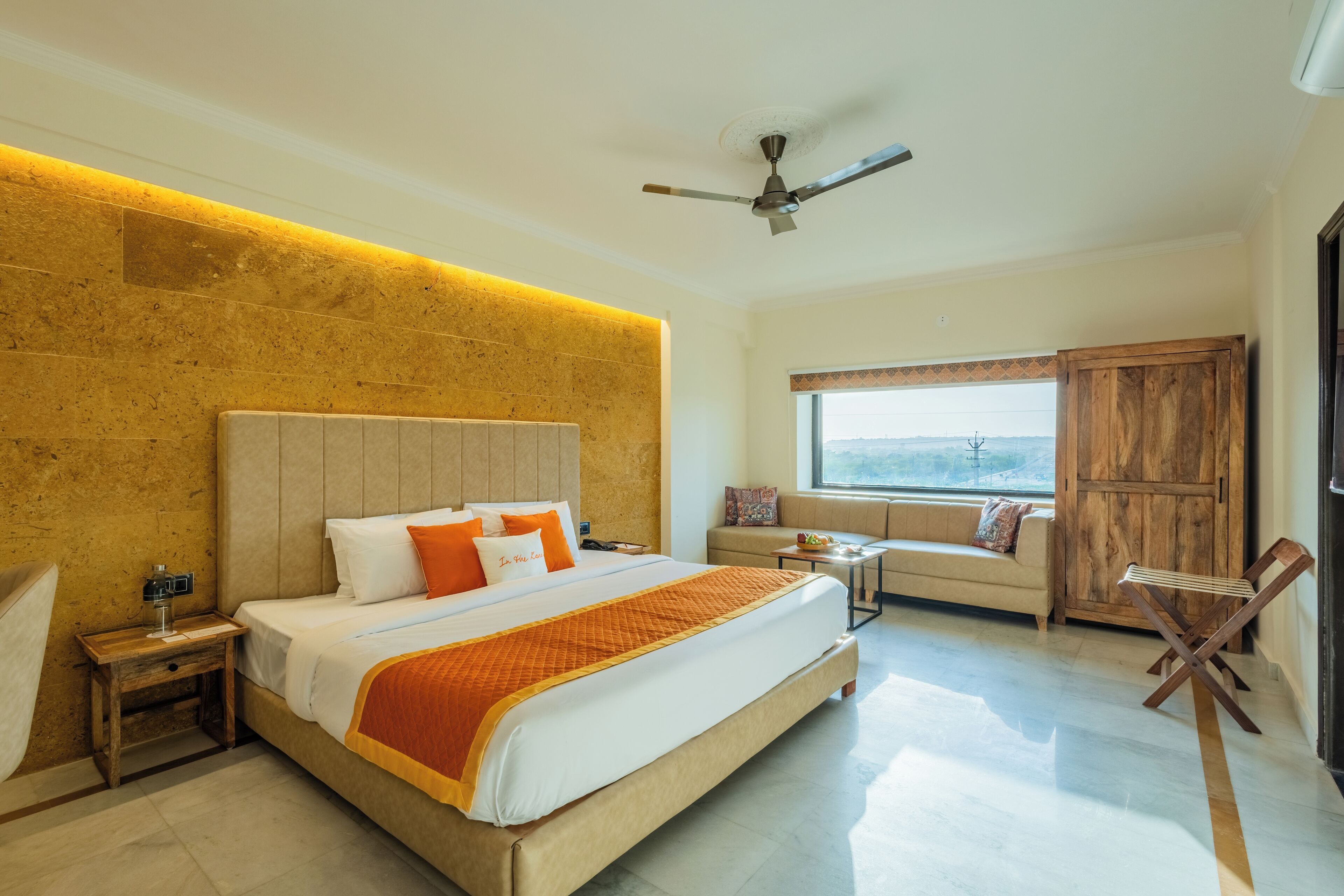 Une chambre d’hôtel moderne avec un grand lit, une tête de lit en bois, un ventilateur de plafond, une fenêtre avec vue, une armoire en bois et une petite table avec une chaise.