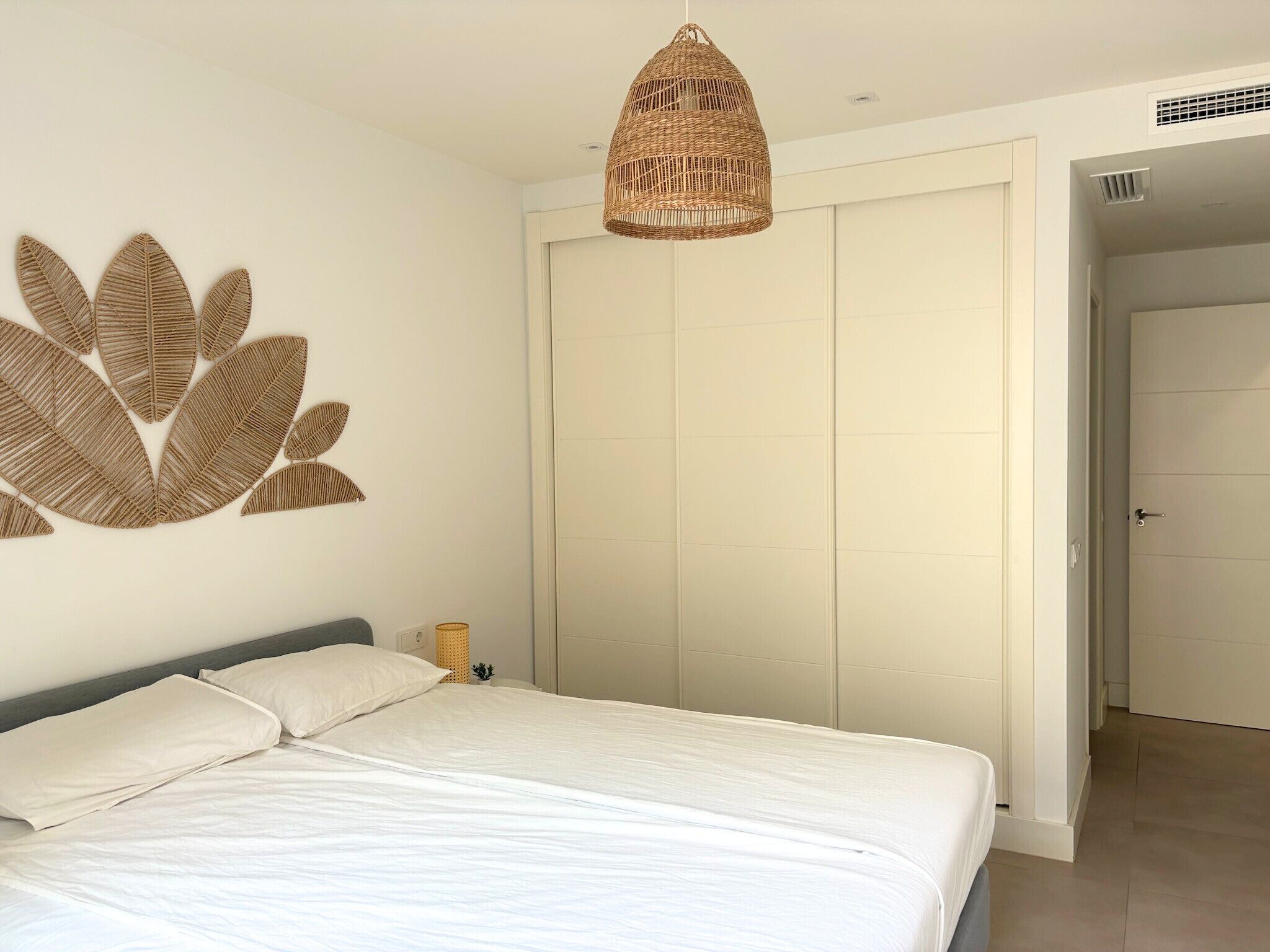 A bedroom with a bed, a wall-mounted decorative leaf pattern, a woven pendant light, and a closed wardrobe.