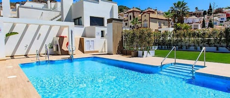 A swimming pool with a clear blue water, surrounded by a white building with a modern design, and a well-maintained lawn area.