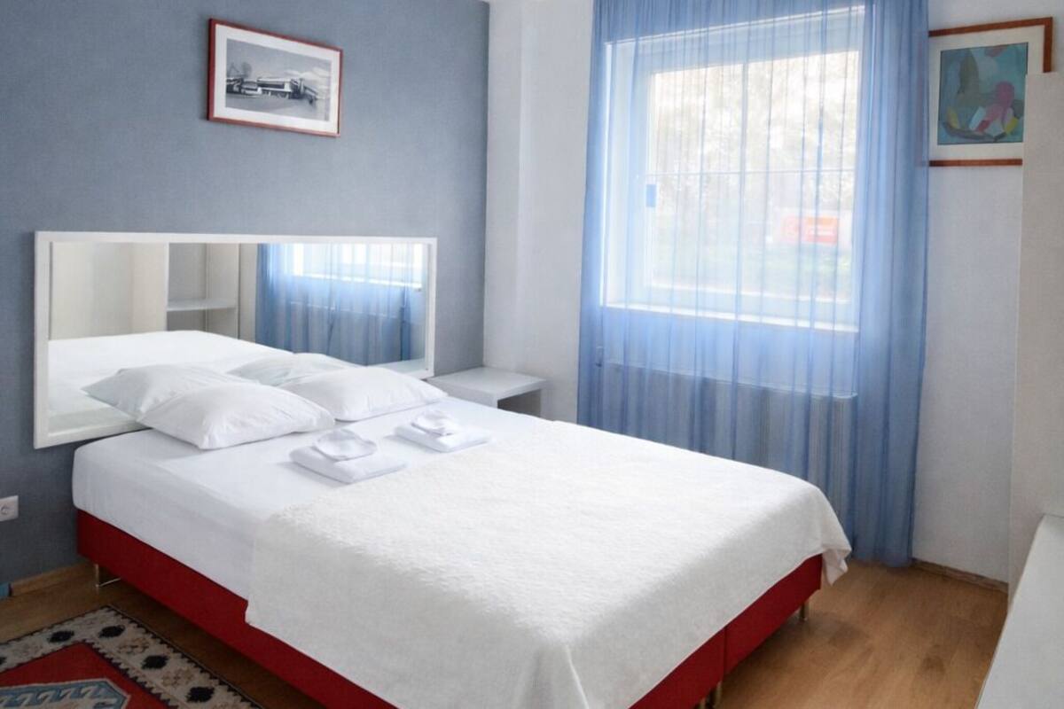 A hotel room with a bed, a nightstand, a desk, a chair, a window with curtains, and a ceiling light.