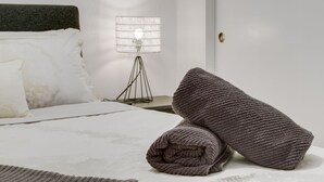 Un letto rifatto con lenzuola bianche, una testiera scura, una lampada da terra moderna e una coperta arrotolata sul letto.