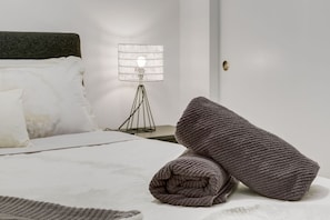 En snyggt bäddad säng med vita sängkläder, en mörk sänggavel, en modern golvlampa och en hoprullad filt på sängen.