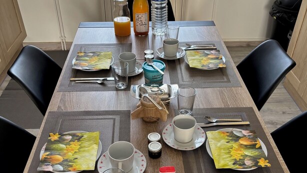 Een eettafel gedekt met borden, bekers en bestek, met een fles sap en een thermoskan erop.