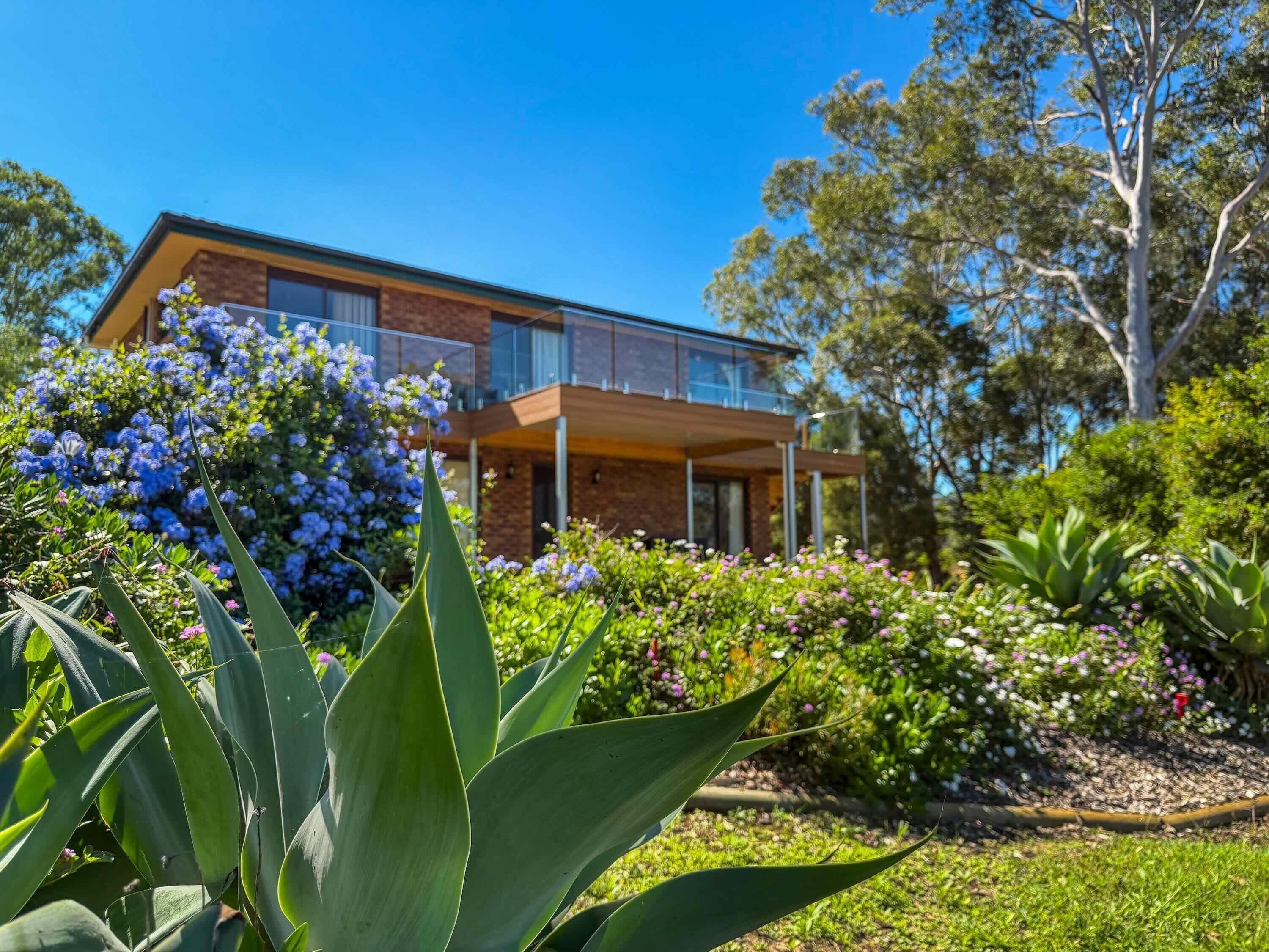 A two-story house with a balcony, surrounded by lush greenery and flowering plants.