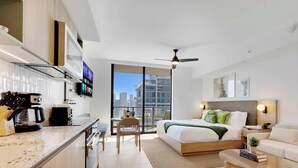 A modern hotel room with a bed, a dining area, a kitchen, and a large window offering a city view. - Oasis Studio with Gym & Pool (Miami)