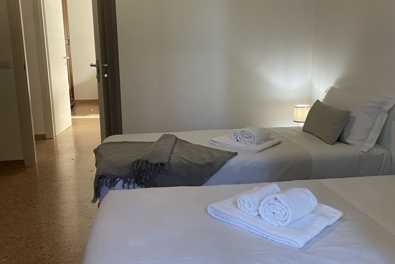 Ein Hotelzimmer mit zwei Betten, jedes mit weißer Bettwäsche und einem gefalteten Handtuch. An einem Bett befindet sich ein Nachttisch mit einer Lampe und einem Kopfkissen.