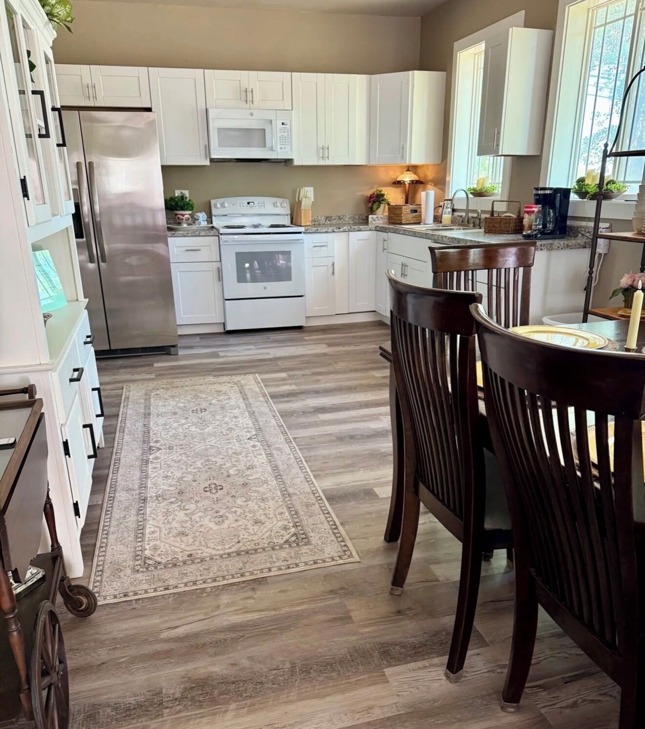 A kitchen with white cabinets, a stainless steel refrigerator, a microwave, an oven, and a dining area with wooden chairs and a rug.