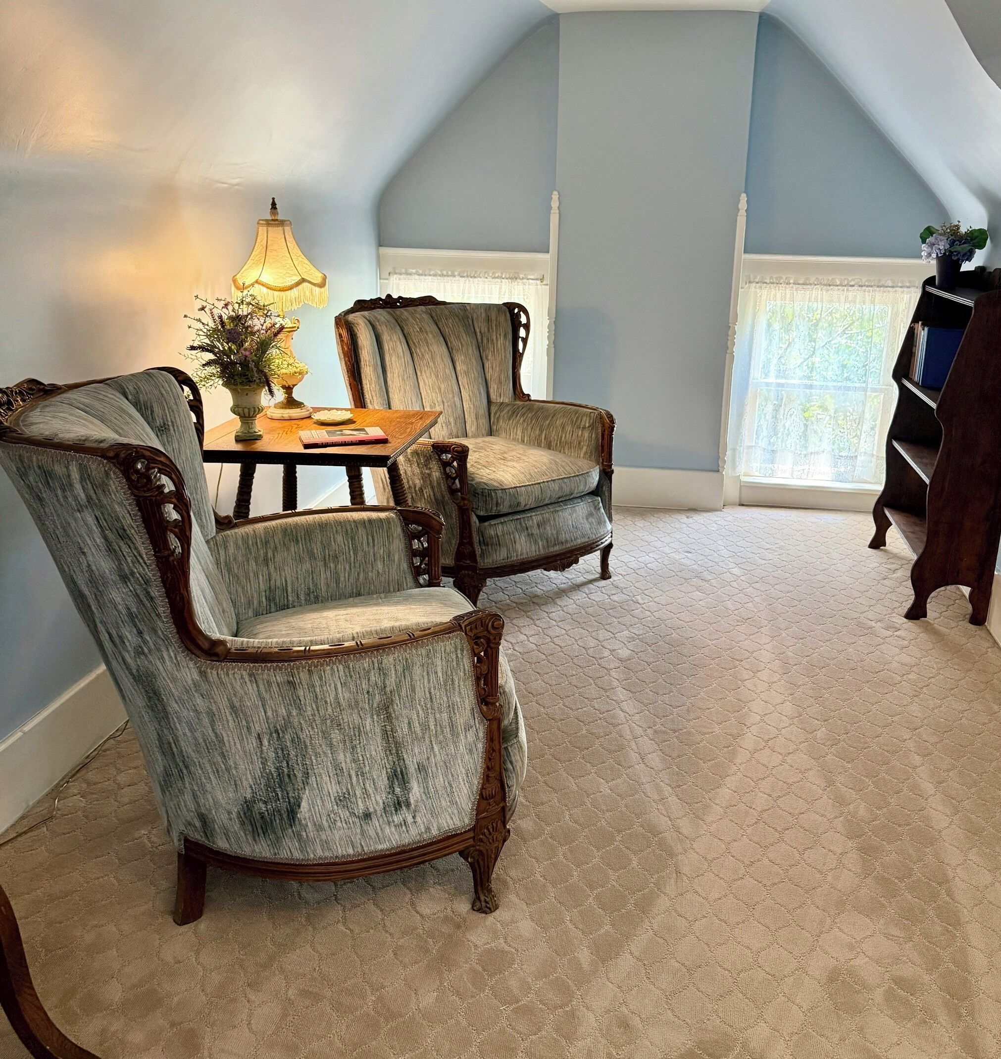 A room with two upholstered chairs, a table with a lamp, and a bookshelf.