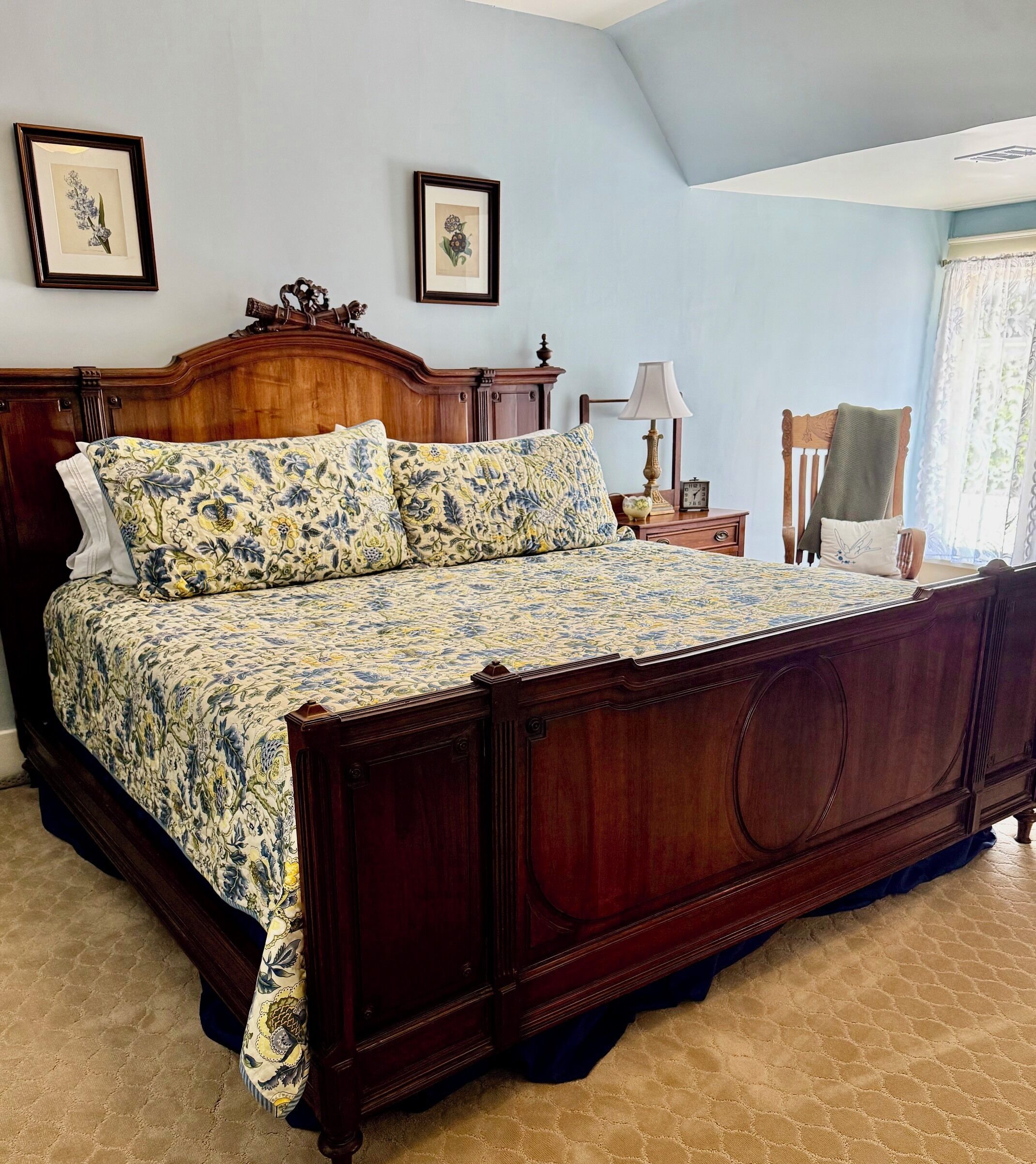 A bedroom with a large bed, wooden headboard, floral bedspread, two framed pictures on the wall, a bedside table with a lamp, and a chair.