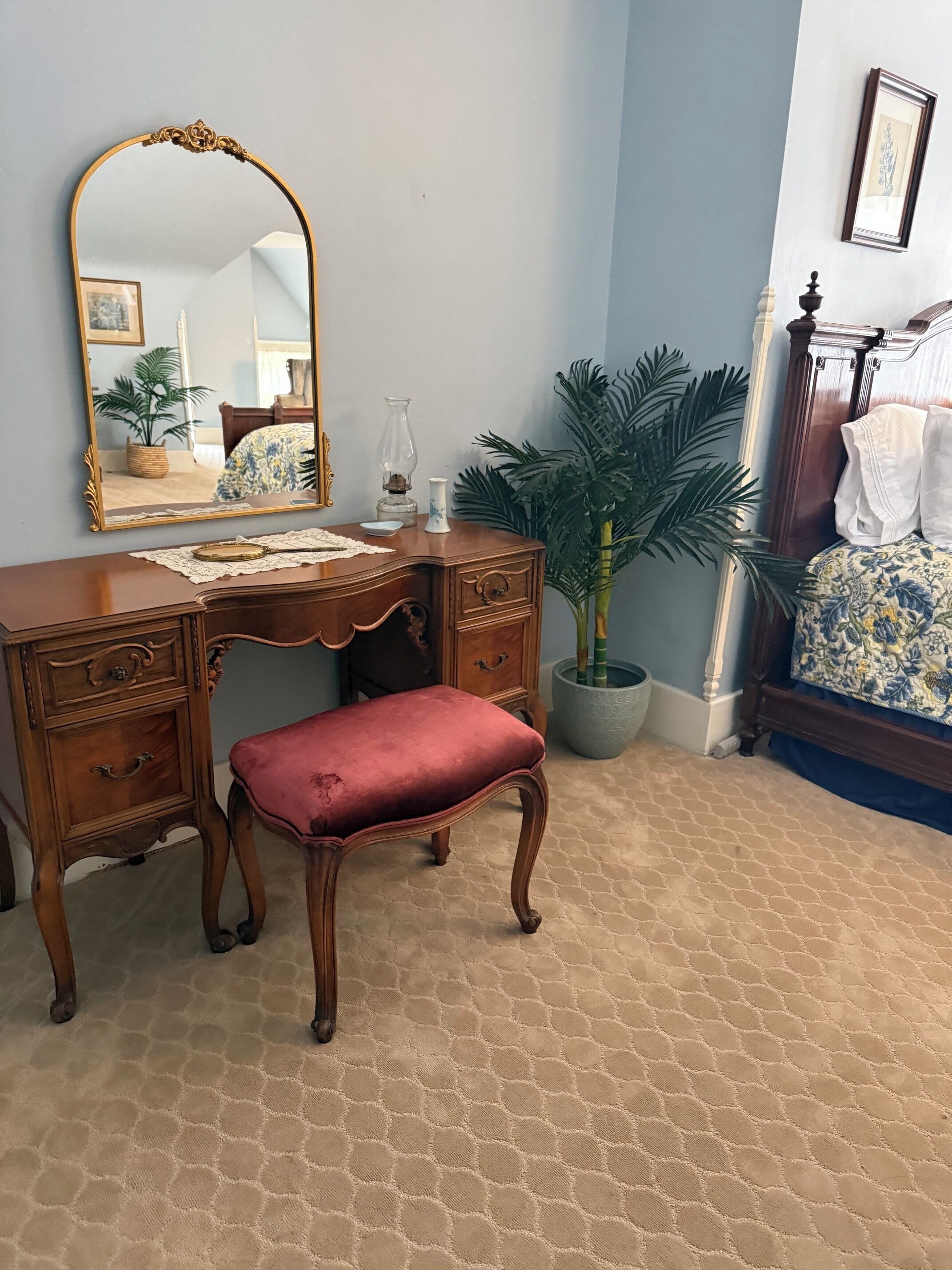 Una habitación con un aparador de madera, un espejo, un banco tapizado rojo, una planta en maceta y una cama con colcha estampada.