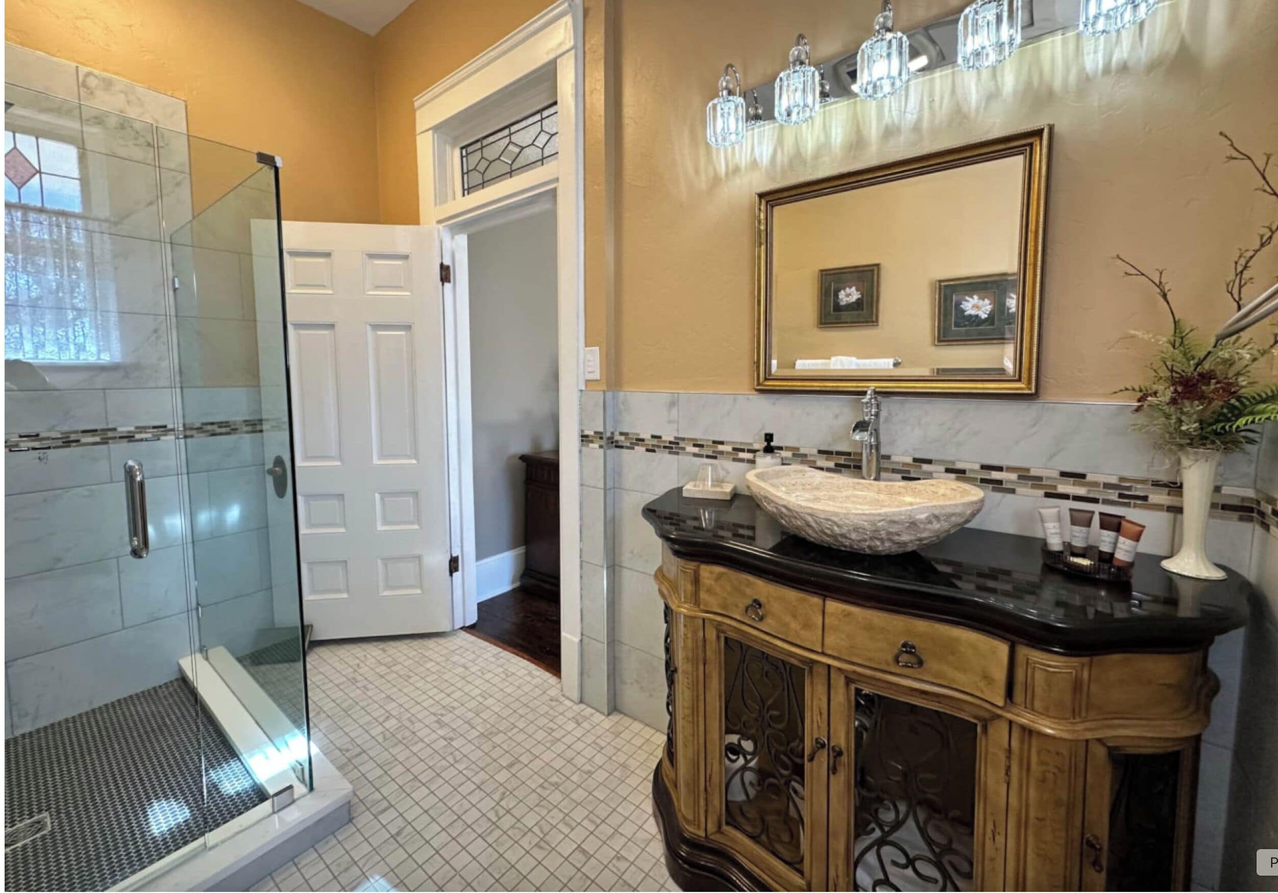 A bathroom with a glass shower, a white door, a vanity with a marble sink, and a mirror with framed pictures.