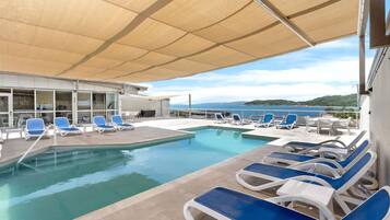Ett poolområde med blå och vita solstolar, en pool och utsikt över havet.