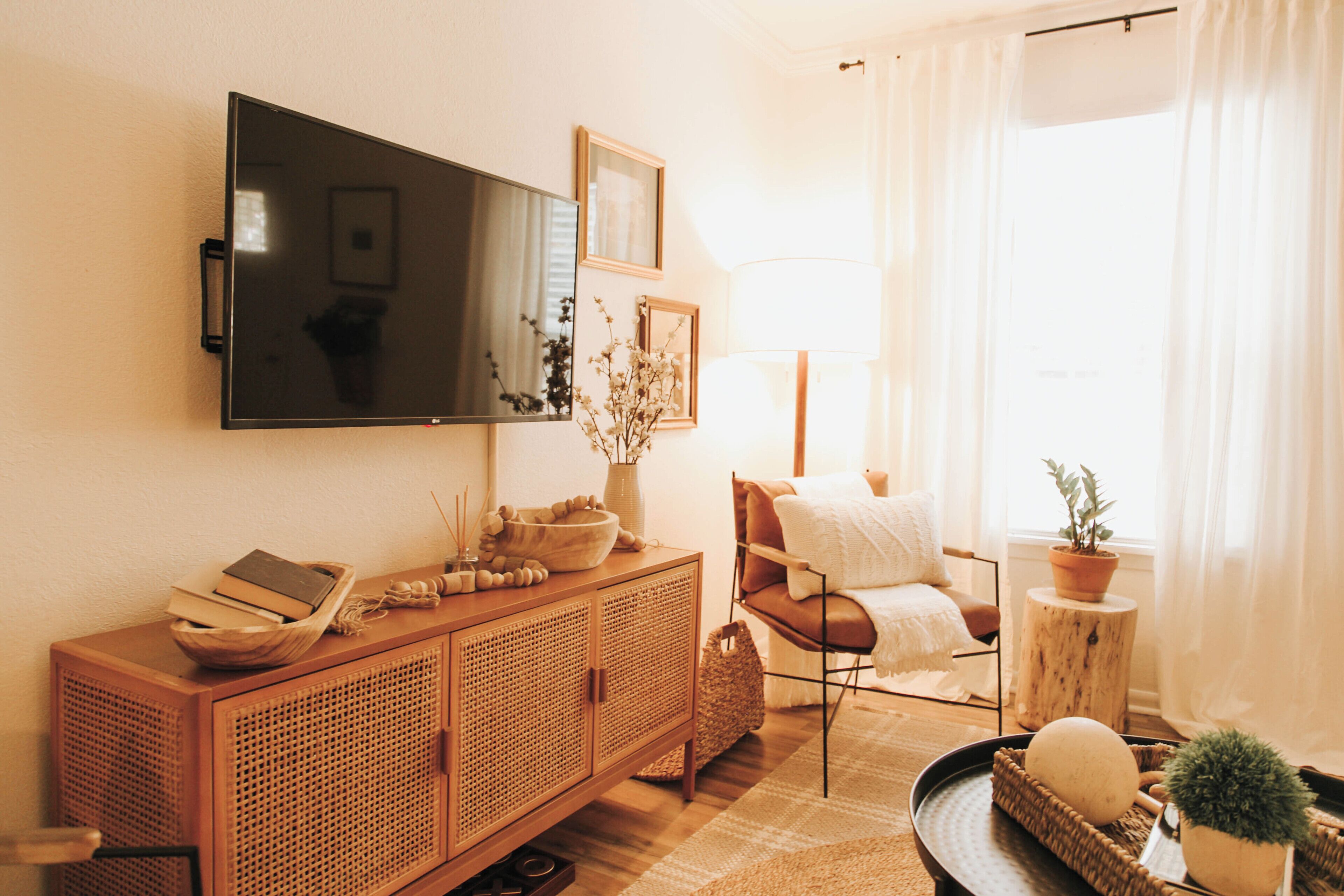 A living room with a wooden cabinet, a television mounted on the wall, a chair, a small table with a plant, and a window with curtains.