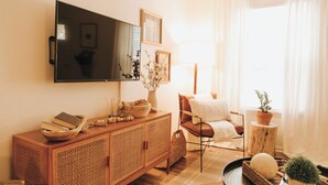 A living room with a wooden cabinet, a television mounted on the wall, a chair, a small table with a plant, and a window with curtains. - Stay & Play at the Charming Woodlawn Cottage (Hot Springs)