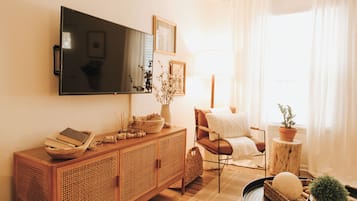 A living room with a wooden cabinet, a television mounted on the wall, a chair, a small table with a plant, and a window with curtains.
