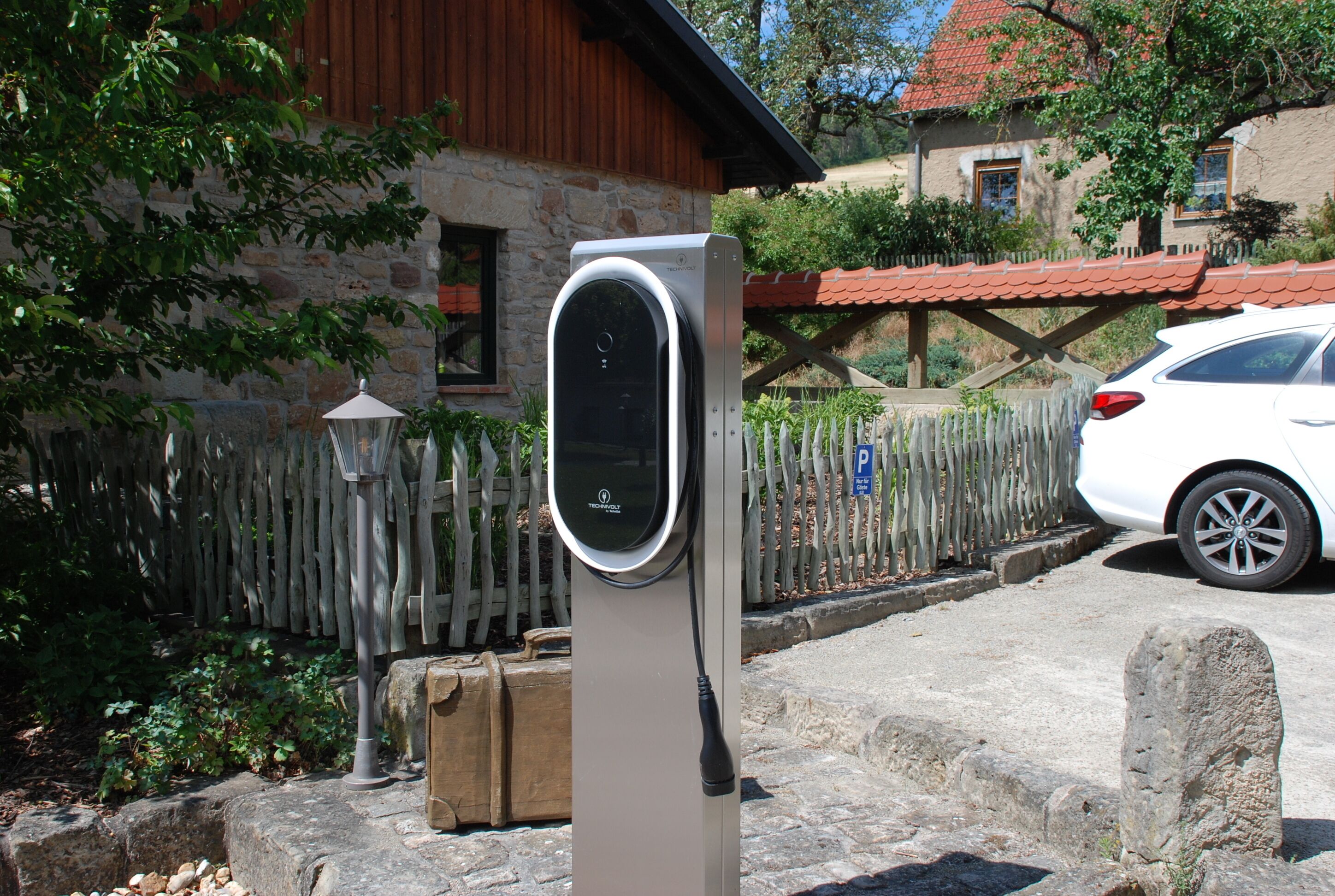A charging station in a residential area with a stone house, a wooden fence, and a parked car.