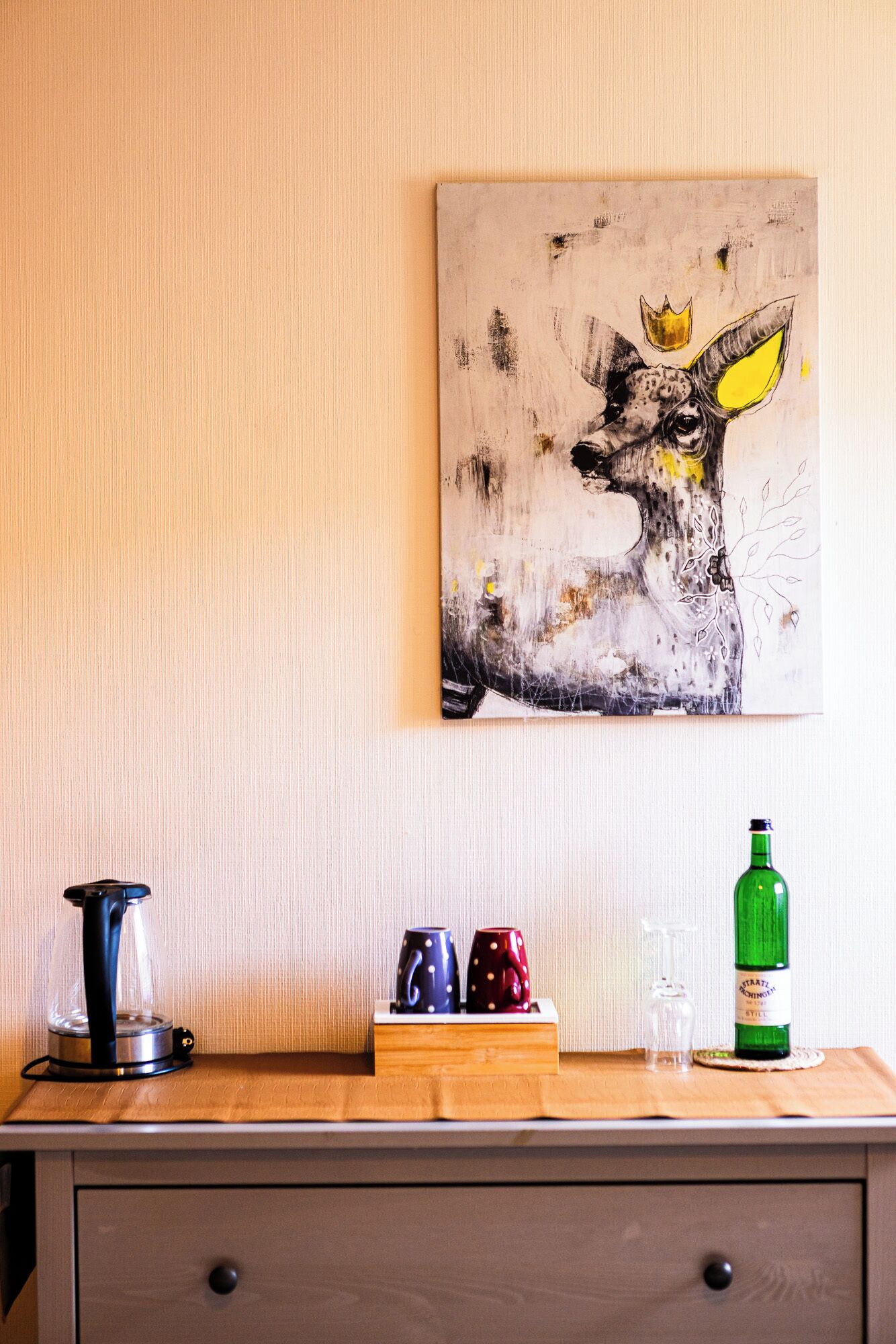 Un mur orné d’un tableau représentant un renard, une machine à café, deux tasses, un verre et une bouteille, le tout posé sur une surface en bois.