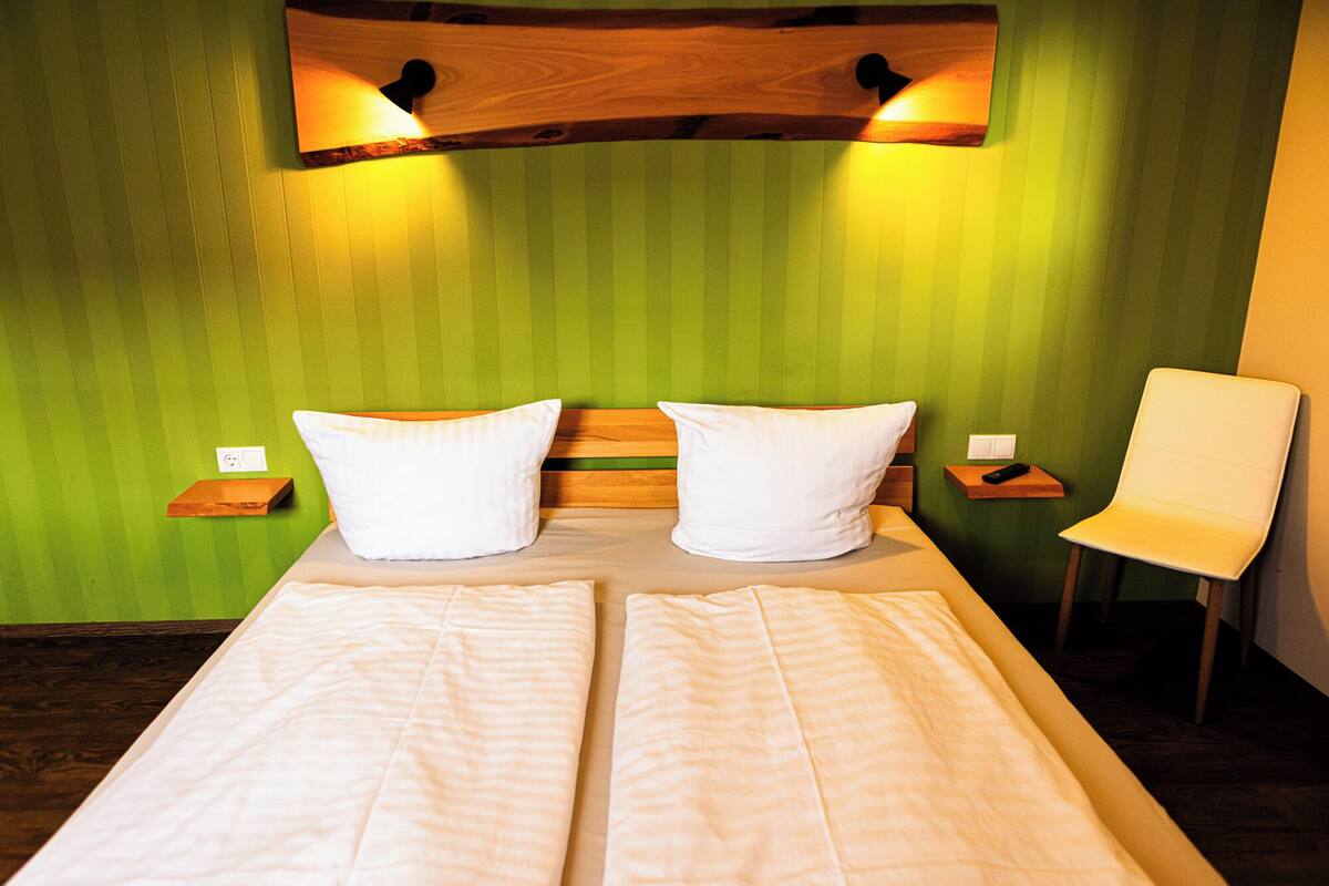 A bed with two pillows, a wooden headboard, a nightstand with a remote, and a chair.