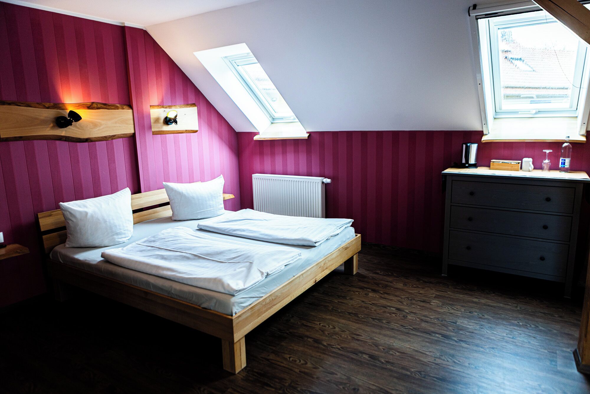 Un dormitorio con cama de madera, ropa de cama blanca, un aparador y dos tragaluces.