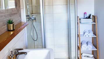 Une salle de bain moderne avec une douche à parois de verre, un lavabo blanc doté d’un distributeur de savon en bois et une étagère en bois où sont posées des serviettes.