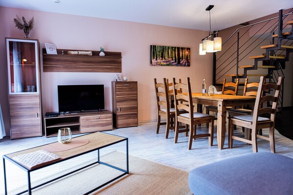 Una sala moderna con una televisión de pantalla plana, muebles de madera, una mesa de comedor con sillas, una escalera y un cuadro en la pared.