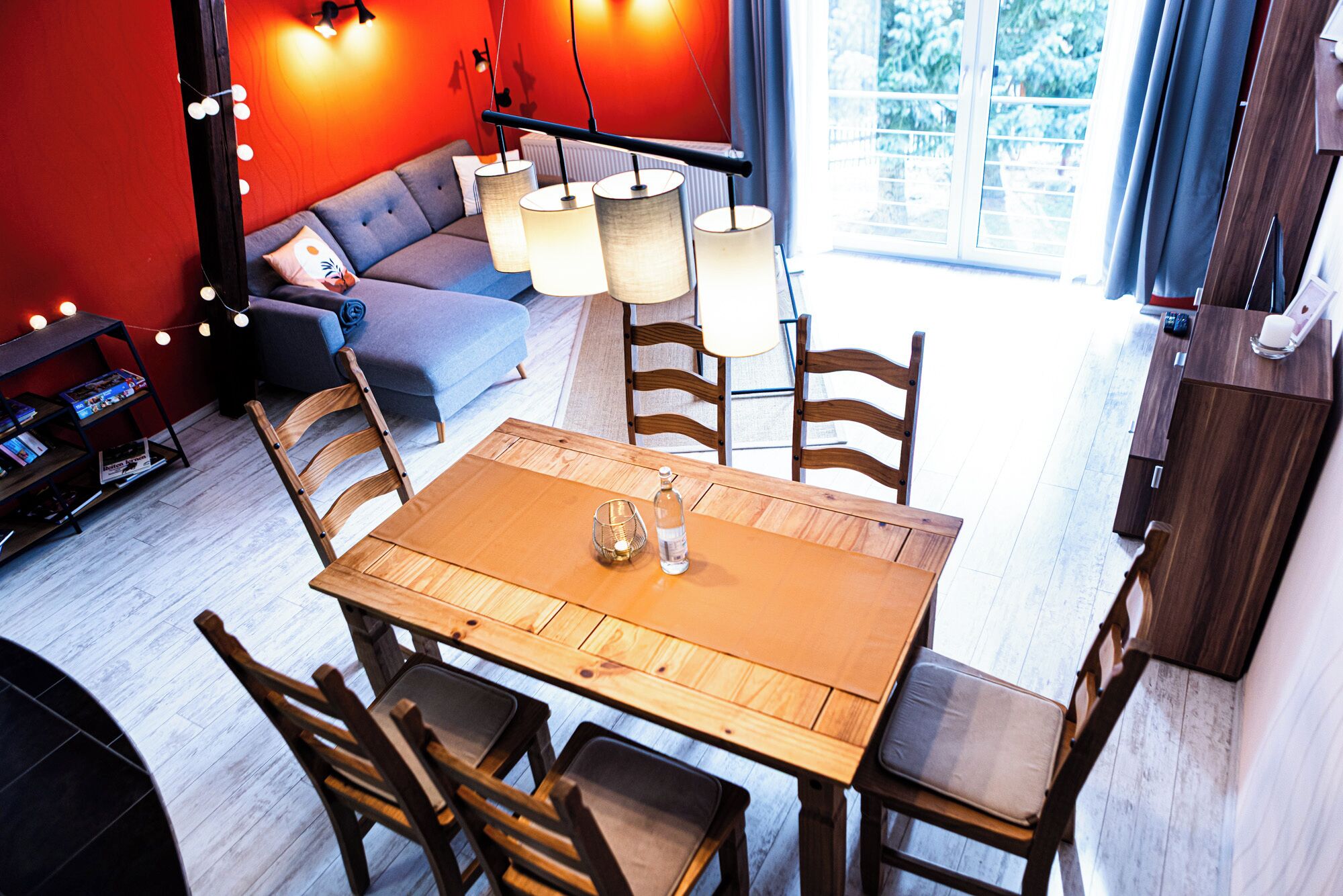 Ein modernes Wohnzimmer mit einem Holzesstisch, Stühlen, einer Couch und einem Couchtisch. Im Raum befindet sich ein Bücherregal und eine Lampe.