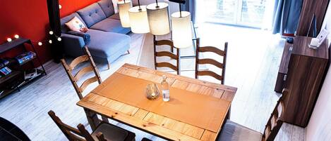 Ein modernes Wohnzimmer mit einem Holzesstisch, Stühlen, einer Couch und einem Couchtisch. Im Raum befindet sich ein Bücherregal und eine Lampe.