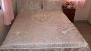 A bedroom with a bed, a nightstand, and a window with curtains. - Villa Nickerie (Nieuw Nickerie)